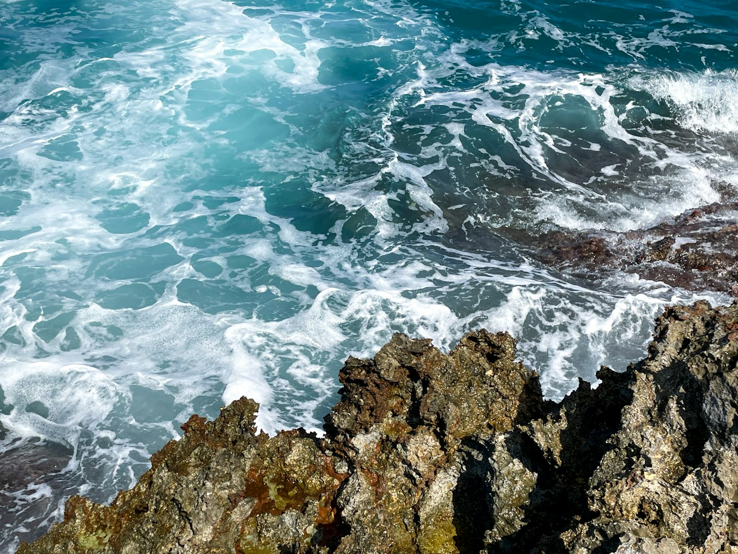 Olas turquesas del océano rompen contra rocas negras y dentadas.